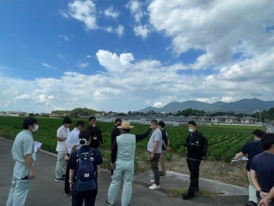 大豆ほ場見学のようす。大豆ほ場の前でJAたがわ麦大豆部会の山口部会長が話しているのを参加者が聞いている。